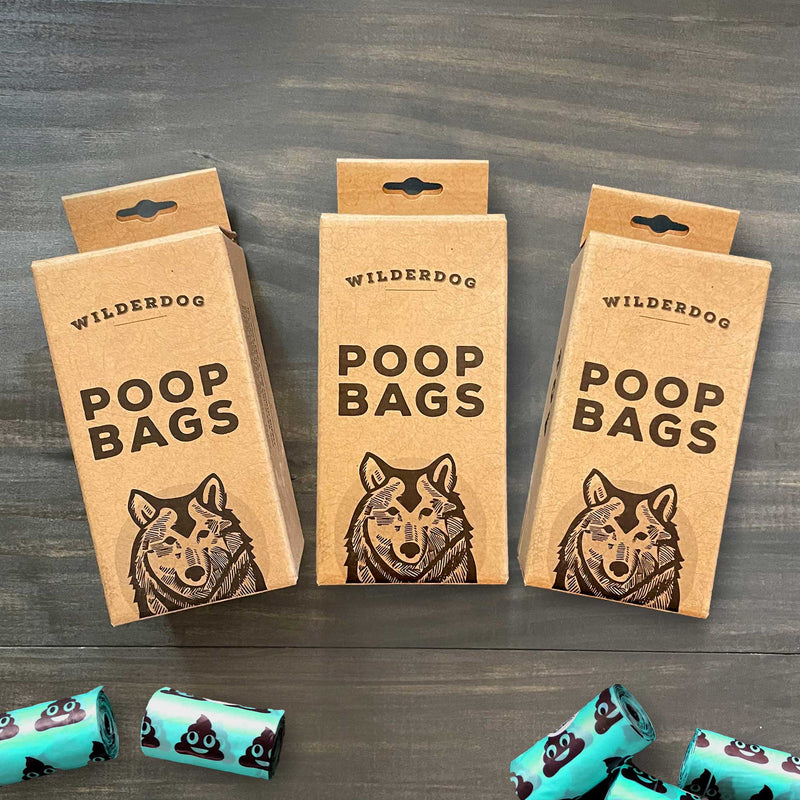 Three boxes of Dog Poop Bags - 3 Pack, each with a wolf illustration, are displayed on a wooden surface along with teal extra-thick bags decorated with playful poop emojis.